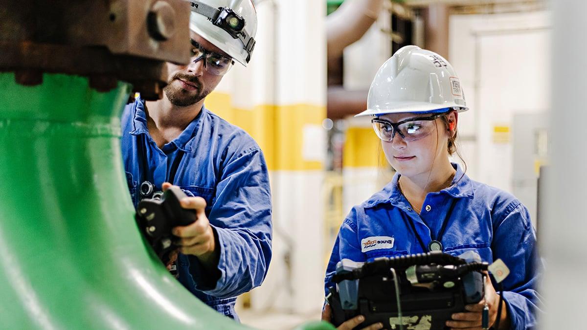 Woman and man, wearing a hardhat and safety goggles, inspecting equipment.